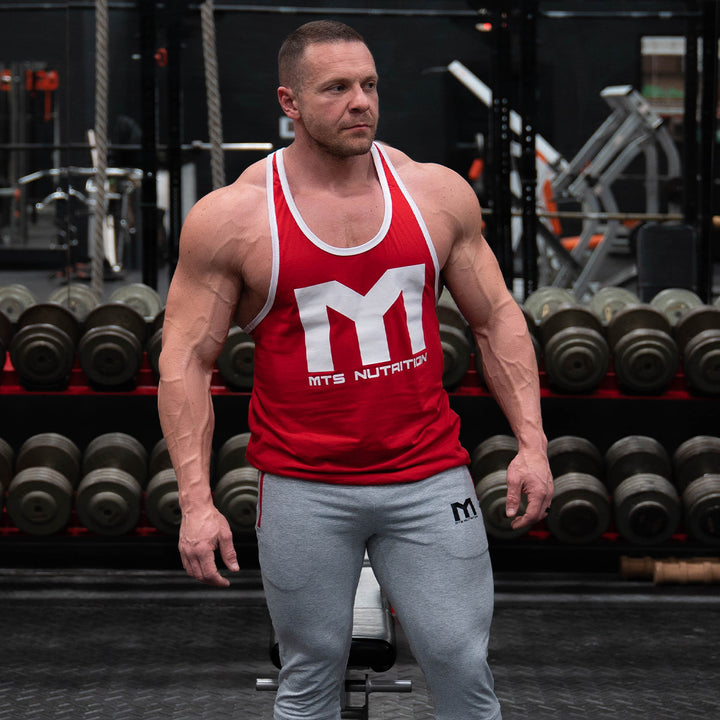 Marc Lobliner stands facing the camera in a front physique pose in a gym, showing developed chest, shoulders and arms.