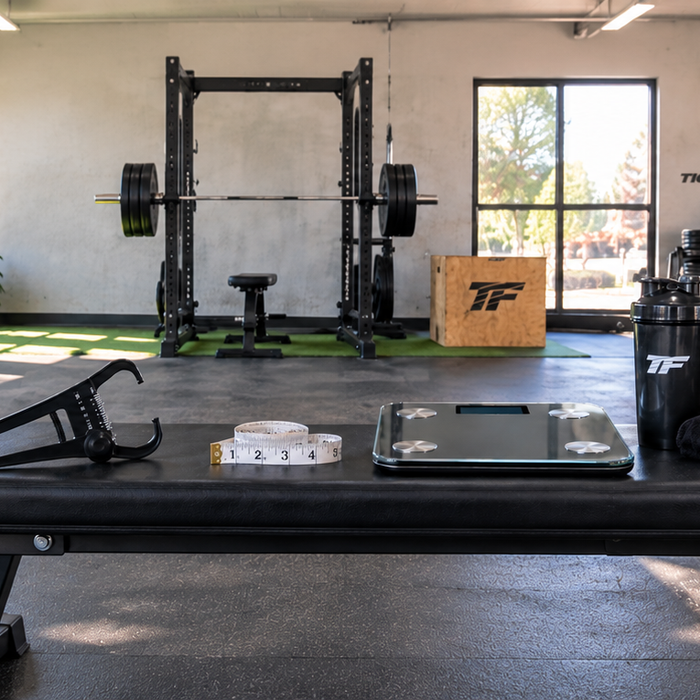 Body fat measurement tools including skinfold calipers, measuring tape, and a digital scale displayed on a bench in a bright Tiger Fitness gym.