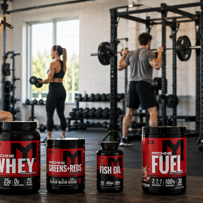MTS Nutrition supplement stack featuring Machine Whey, Greens + Reds, Fish Oil, and Machine Fuel in a bright gym with athletes training in the background.