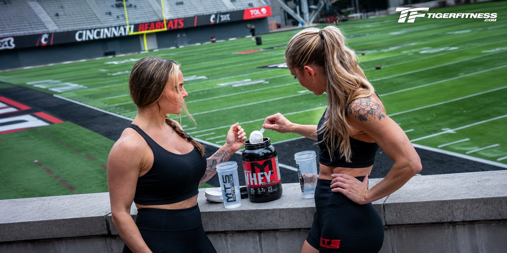 Two female athletes preparing MTS Nutrition whey protein shakes overlooking a football field at a stadium.