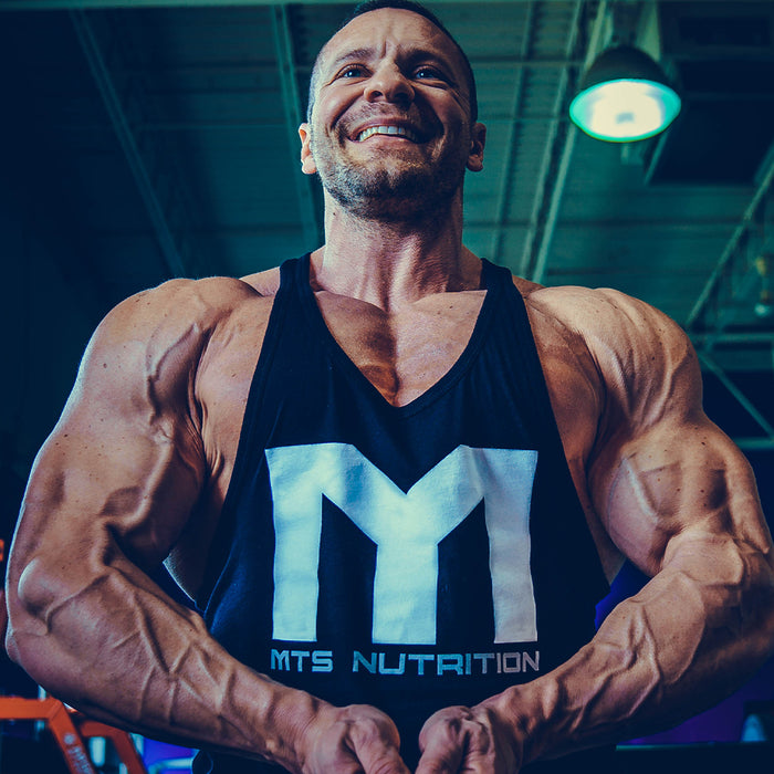 Marc Lobliner in a standing front physique pose, arms hanging at his sides with triceps and forearms visible.