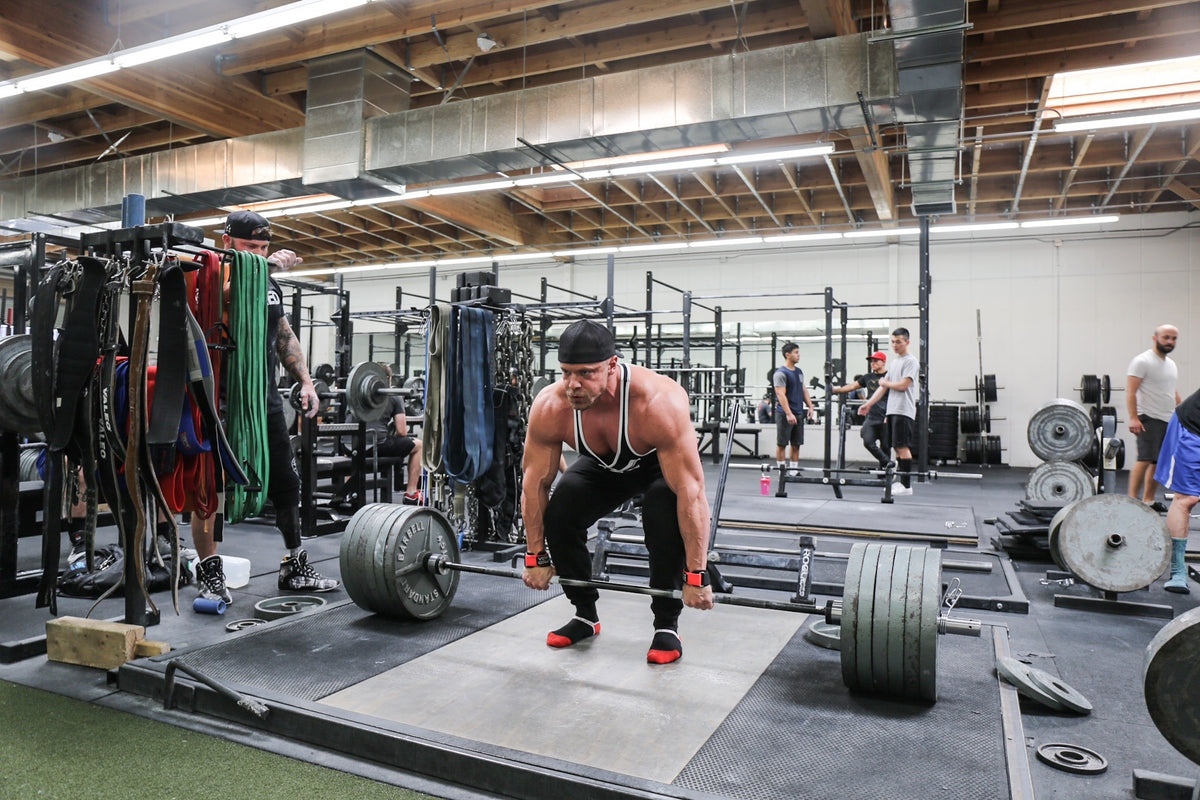 Marc Lobliner locking out a heavy barbell deadlift on a gym platform, bar against the thighs and hips fully extended