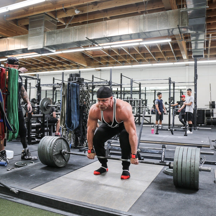 Marc Lobliner locking out a heavy barbell deadlift on a gym platform, bar against the thighs and hips fully extended