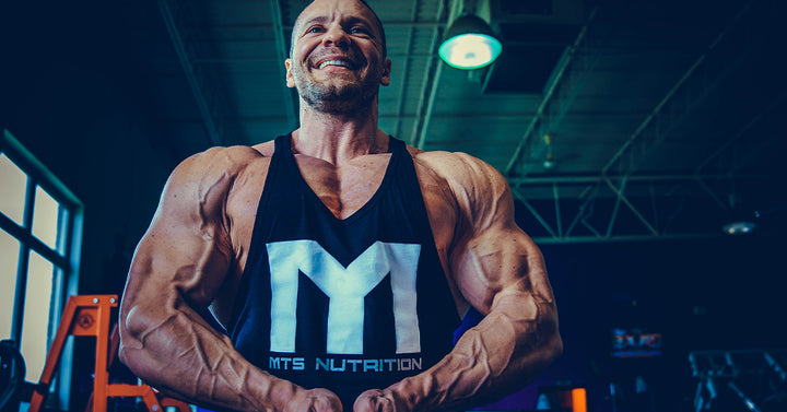 Marc Lobliner standing in a gym in a front physique pose, legs and torso in frame, no machine in the shot.