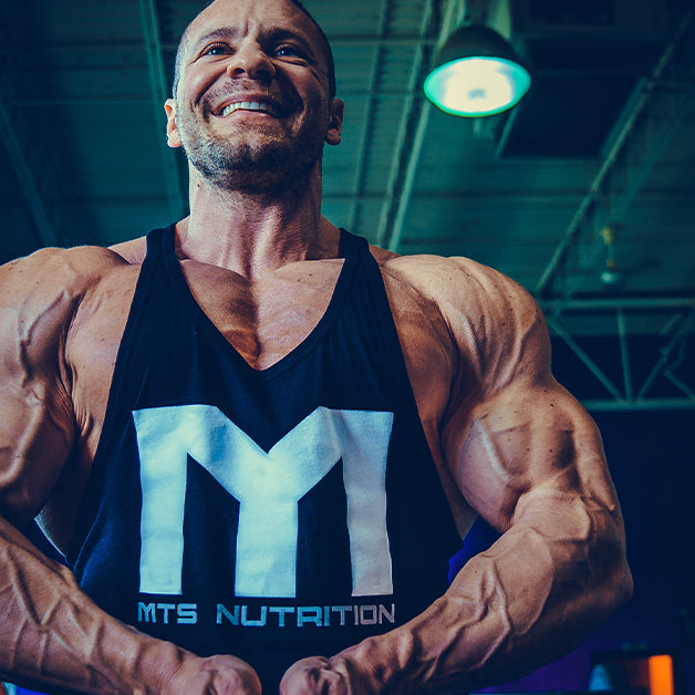 Marc Lobliner standing in a gym in a front physique pose, legs and torso in frame, no machine in the shot.