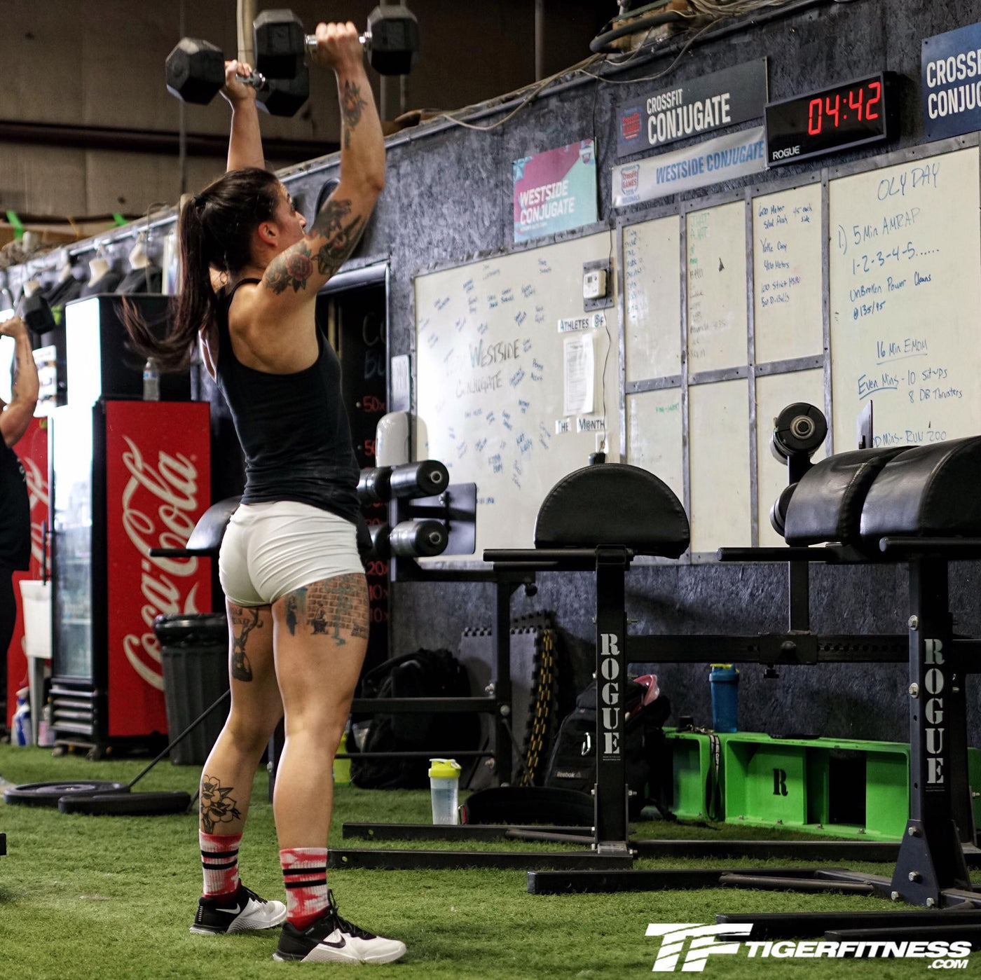 Tattooed male athlete driving a pair of dumbbells overhead in a push press during a high frequency training session