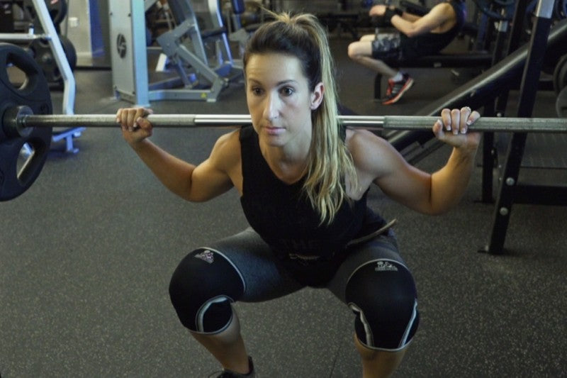 Athlete Kara Corey unracking a loaded barbell for back squats, bar across the upper back and hands set wide