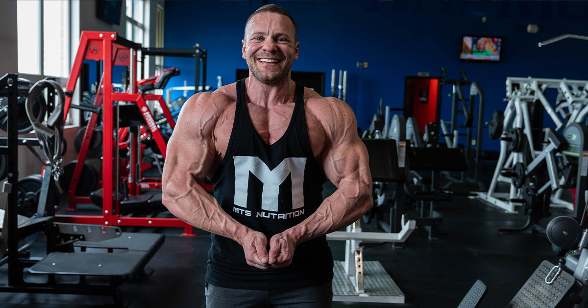 Marc Lobliner in a front physique pose in a gym, shoulders and chest in frame, no bar or bench in the shot.