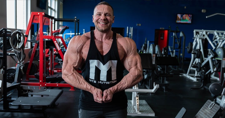 Marc Lobliner in a front physique pose in a gym, shoulders and chest in frame, no bar or bench in the shot.