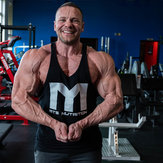 Marc Lobliner in a front physique pose in a gym, shoulders and chest in frame, no bar or bench in the shot.