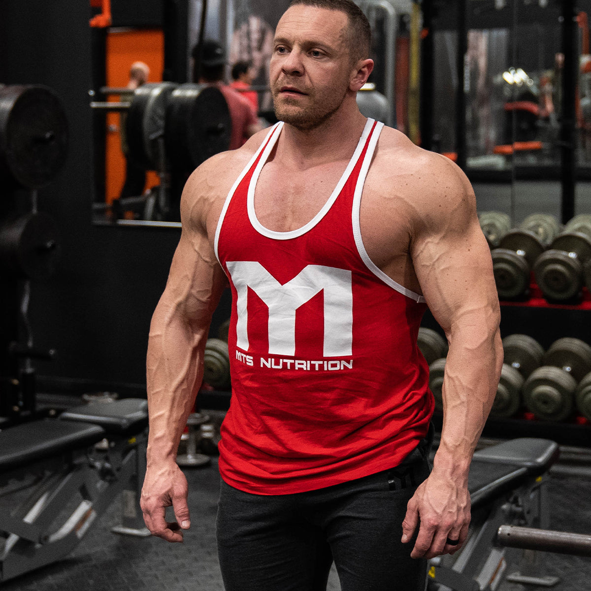 Marc Lobliner standing in a gym in a front physique pose, quads and upper body visible under overhead lighting.