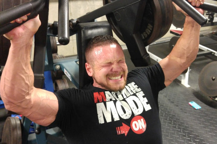 Marc Lobliner seated on a shoulder press machine with his back flat against the pad, pressing the handles overhead.