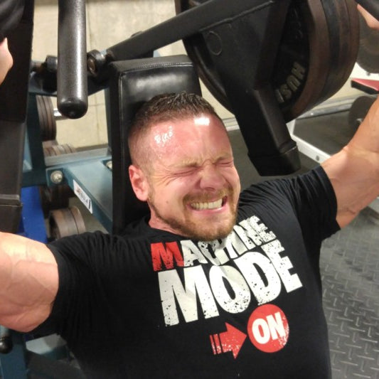 Marc Lobliner seated on a shoulder press machine with his back flat against the pad, pressing the handles overhead.