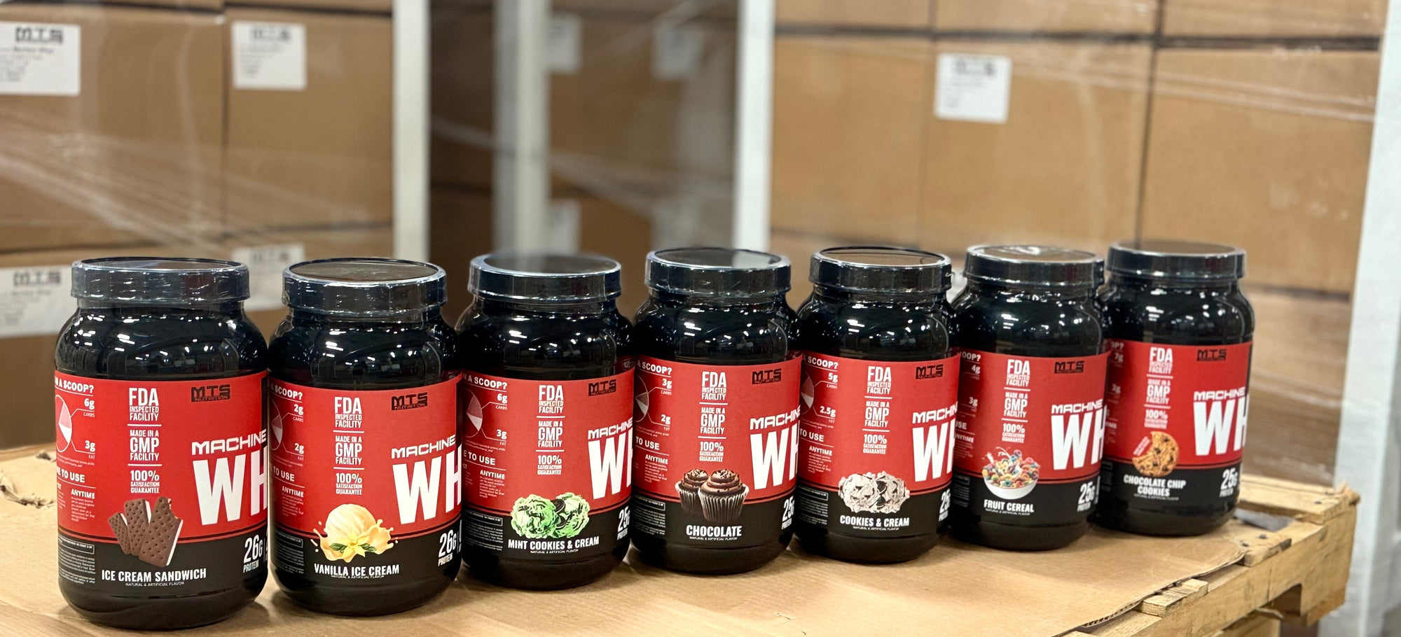Row of MTS Nutrition Machine Whey Protein supplement containers on a pallet with cardboard boxes in the background