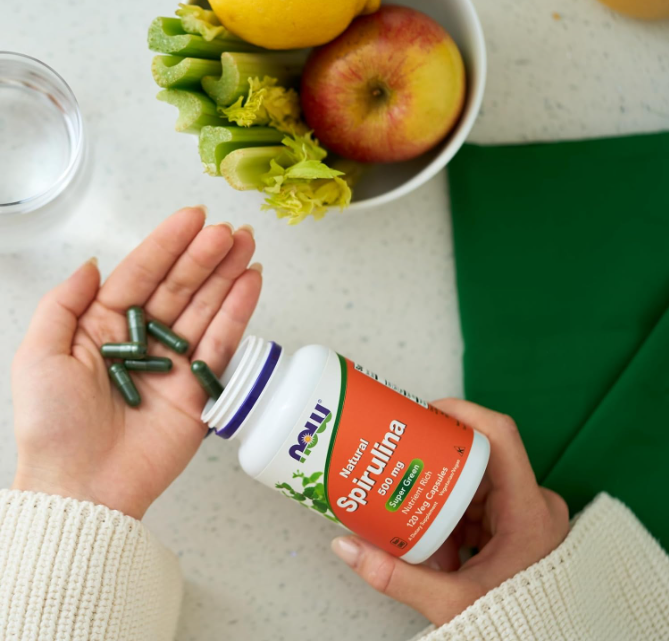 Hand holding NOW Foods Certified Organic Spirulina 500 mg tablets with bottle and fresh fruit on kitchen counter