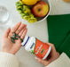Hand holding NOW Foods Certified Organic Spirulina 500 mg tablets with bottle and fresh fruit on kitchen counter